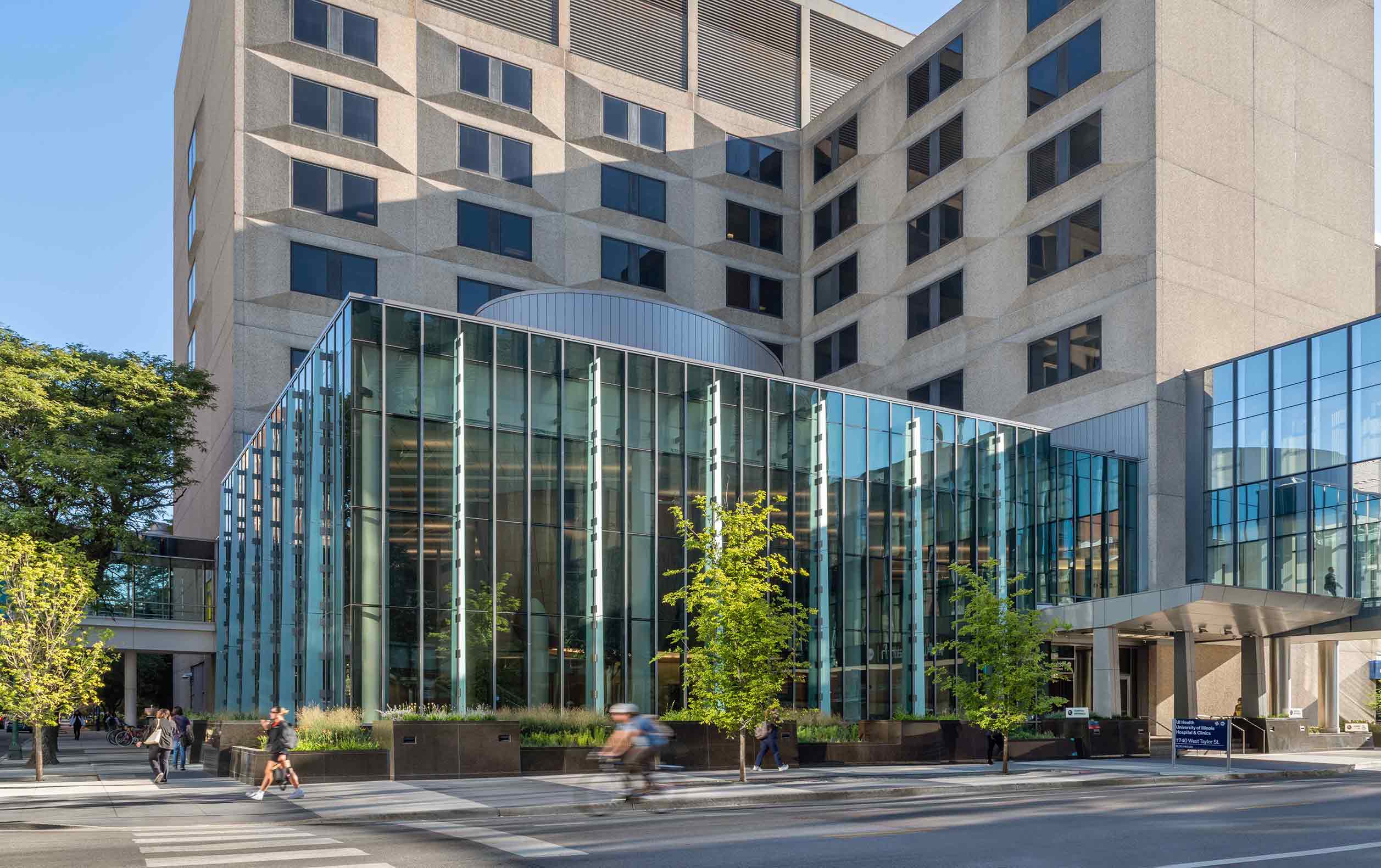 Exterior view of three-story glass building standing before taller concrete structure. An elevated glass pedestrian bridge connects to the glass building.