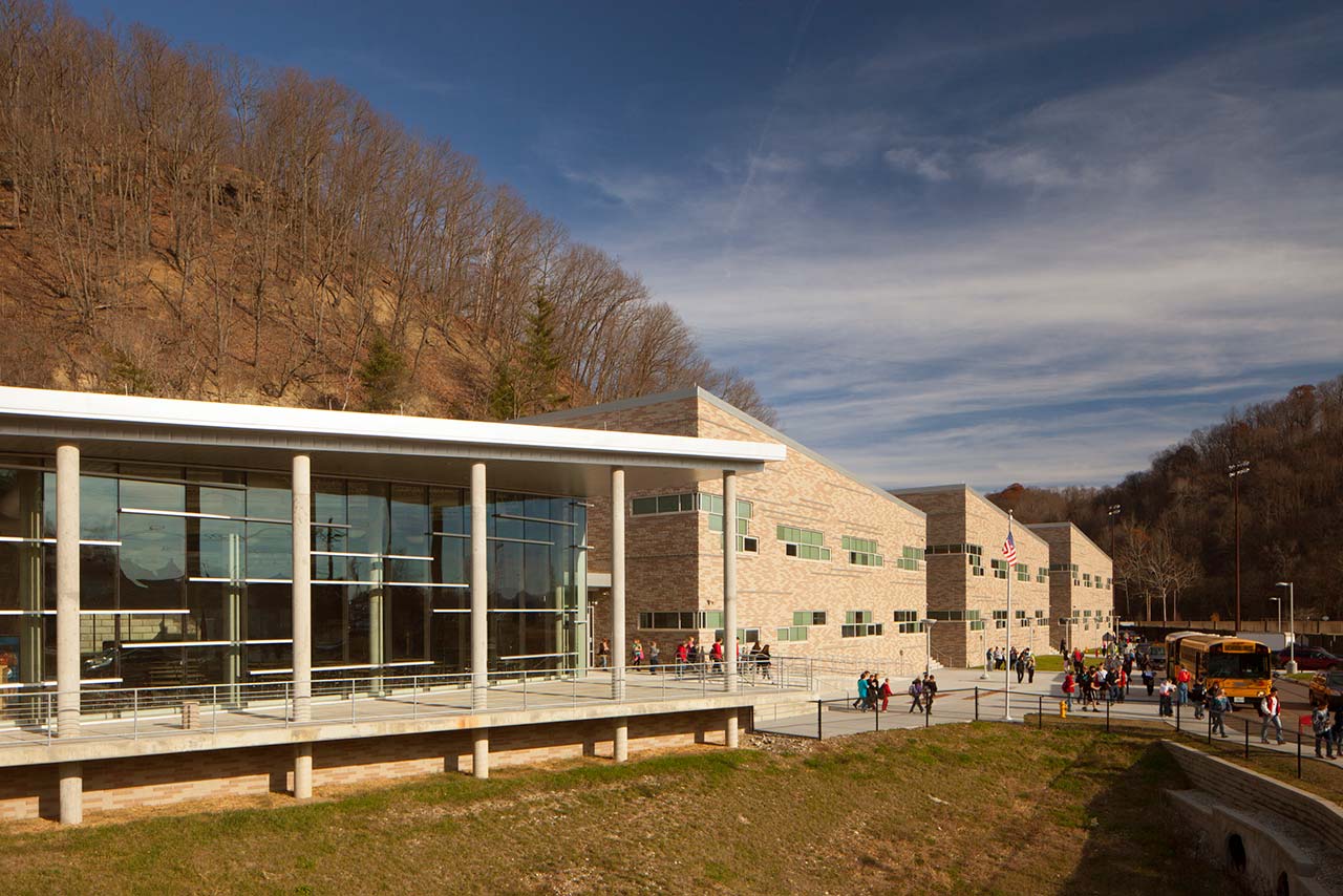 School with large glass wall, stone walls, and tall hill in background.