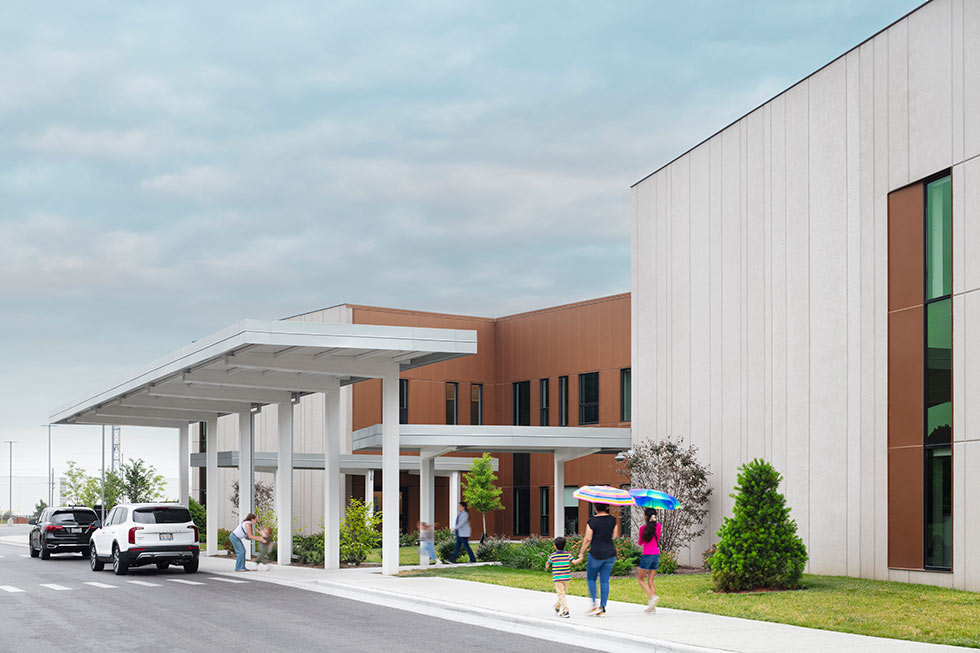 Parents drop off students beneath a metal canopy on a school sidewalk.
