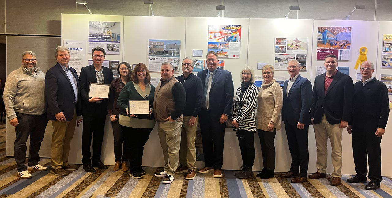 Men and women standing before display of school design project boards and holding award plaques