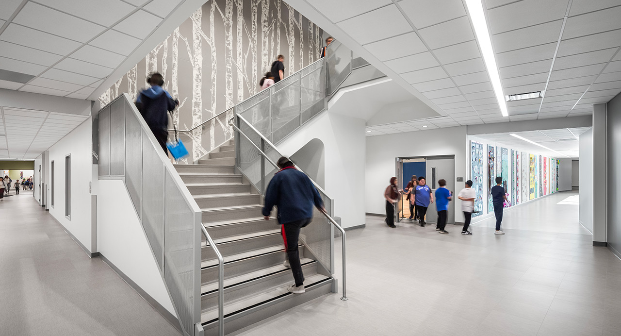 People ascending elementary school staircase with birch tree artwork on wall, students in background